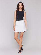 Load image into Gallery viewer, A woman wearing a solid linen skort with a patch pocket on the right side, paired with a black sleeveless top and black strappy sandals.
